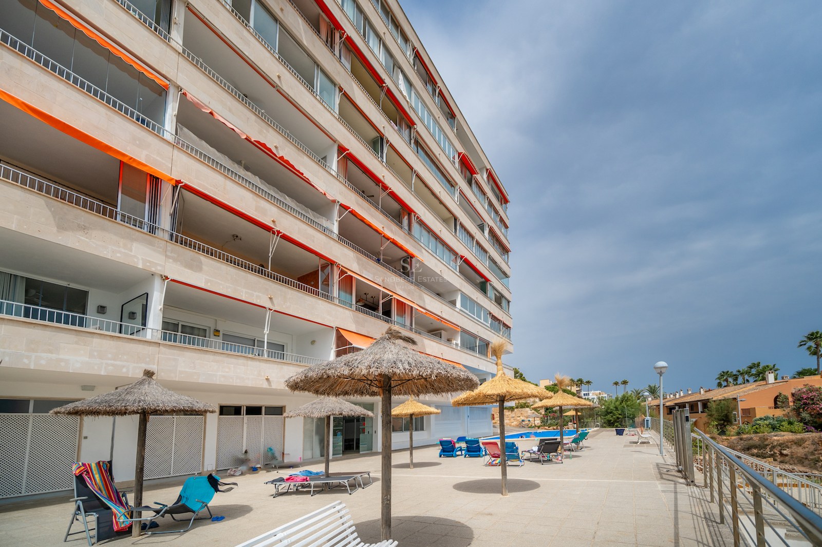Immeuble d'appartements avec balcons, stores orange et espace piscine avec parasols en paille.