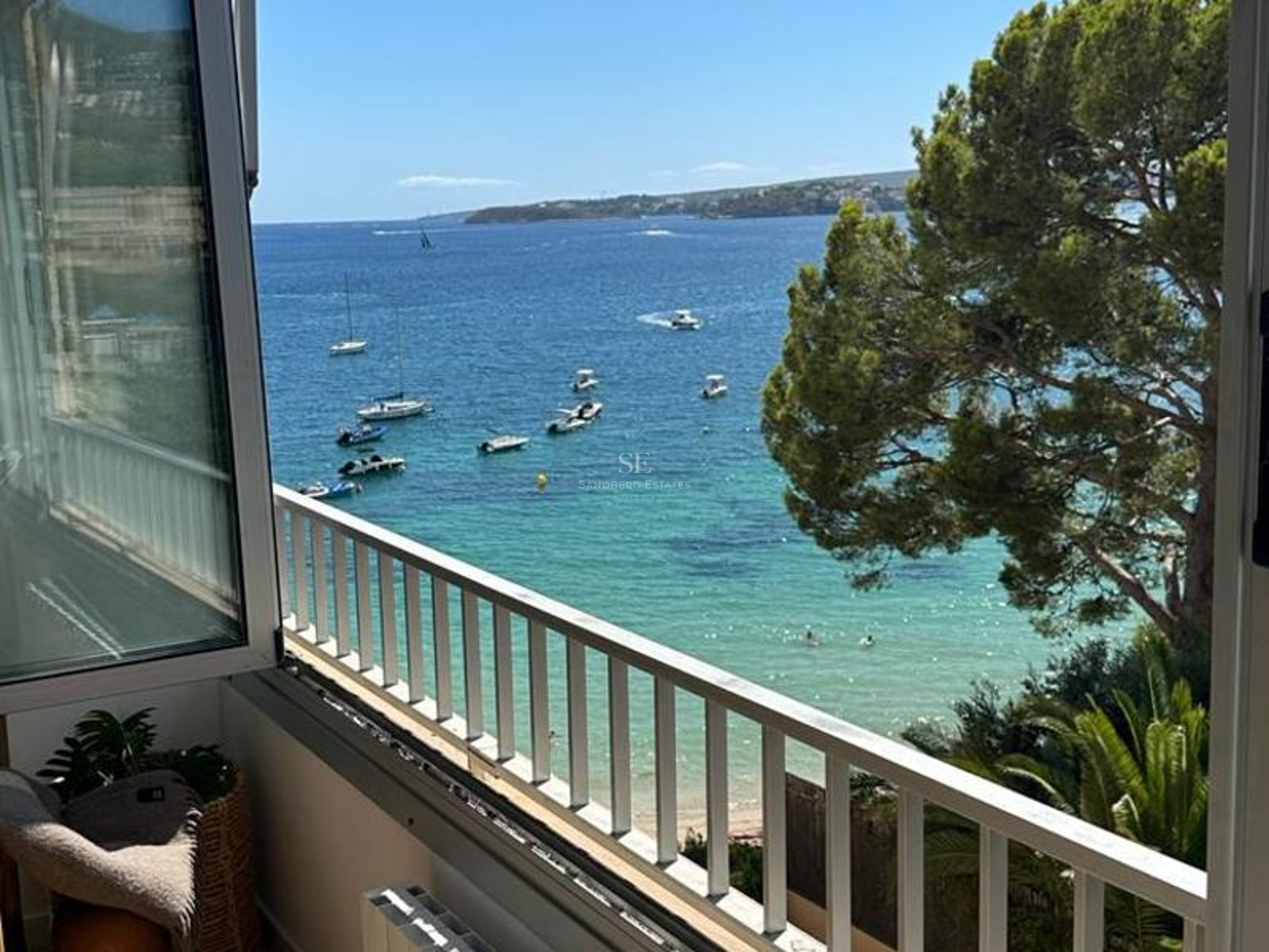 Vue depuis un balcon sur une baie méditerranéenne aux eaux turquoise avec des yachts, un pin et un garde-corps blanc.