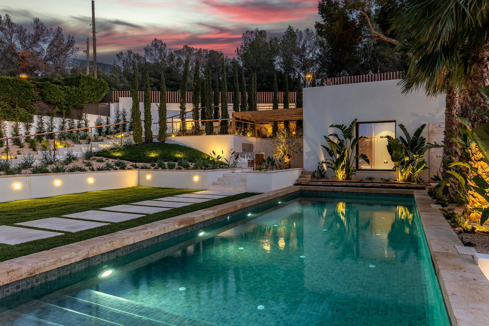 A lit swimming pool with turquoise water at dusk, surrounded by stone paving, green lawns, and tropical plants.