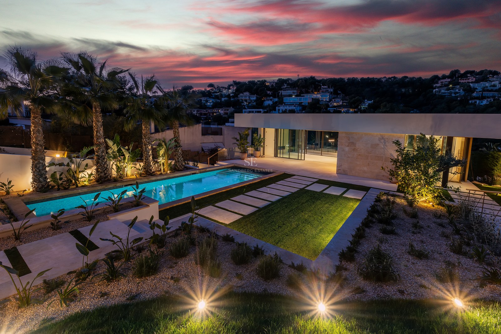 Dusk view of a contemporary villa with an illuminated swimming pool, palm trees, and a dramatic sunset sky.