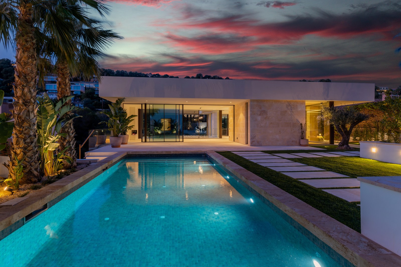 Illuminated swimming pool in front of a modern natural stone villa under a dramatic red sunset sky with palm trees.