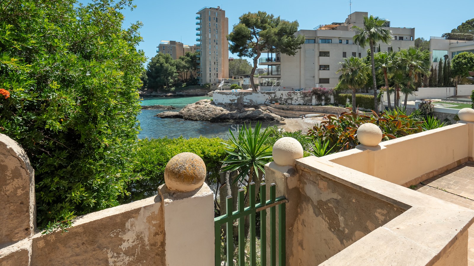 Vue d'une baie aux eaux turquoise et d'un rivage rocheux depuis une terrasse méditerranéenne en pierre.