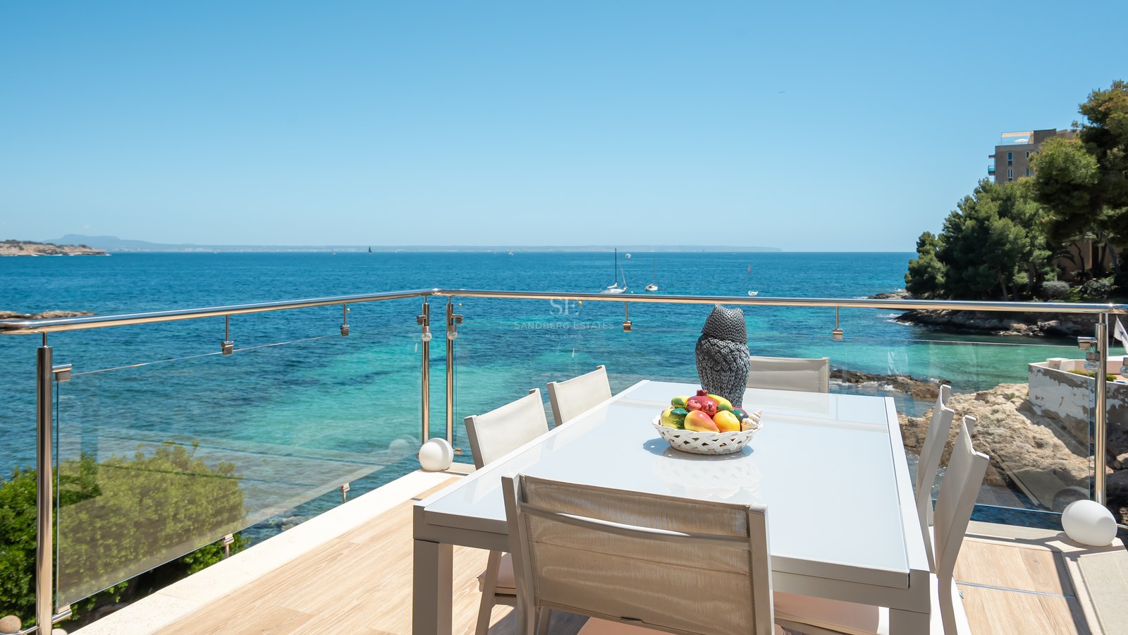 Espace dînatoire extérieur moderne sur une terrasse avec garde-corps en verre surplombant une baie turquoise.