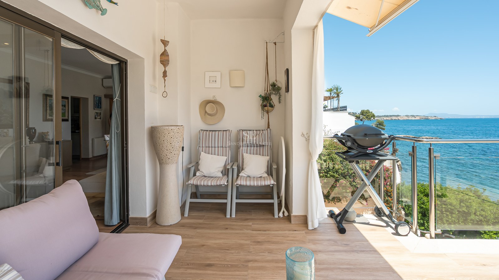 Balcon couvert ensoleillé avec sol imitation bois, coin salon, barbecue et garde-corps en verre surplombant la mer.