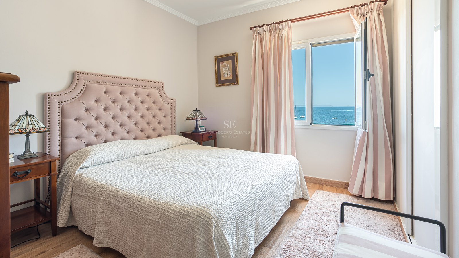 Chambre lumineuse avec tête de lit rose, meubles en bois et fenêtre ouverte donnant sur la mer Méditerranée.