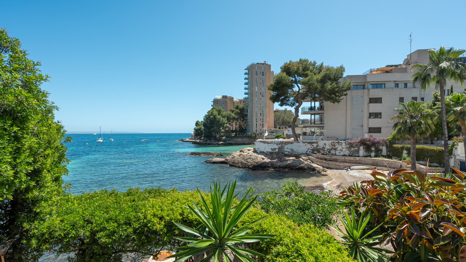 Baie turquoise avec rivages rocheux et bâtiments modernes encadrés par une végétation méditerranéenne luxuriante.