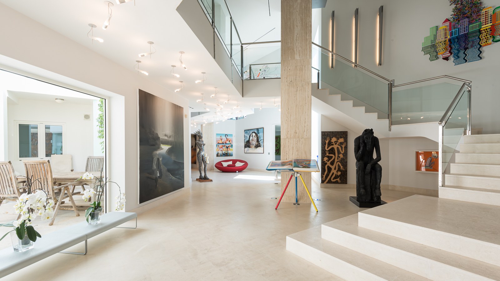 Spacious modern entrance hall with cream travertine floors, glass staircase, and large-scale contemporary artworks.