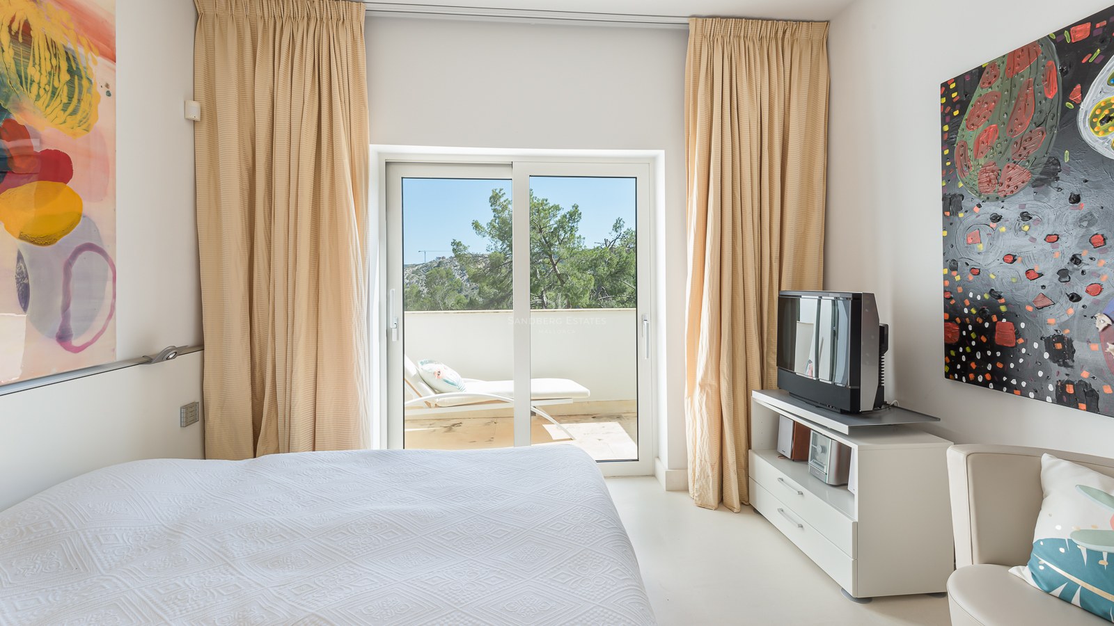 A bright bedroom featuring a white bed, beige curtains, and sliding glass doors leading to a terrace with a lounger.