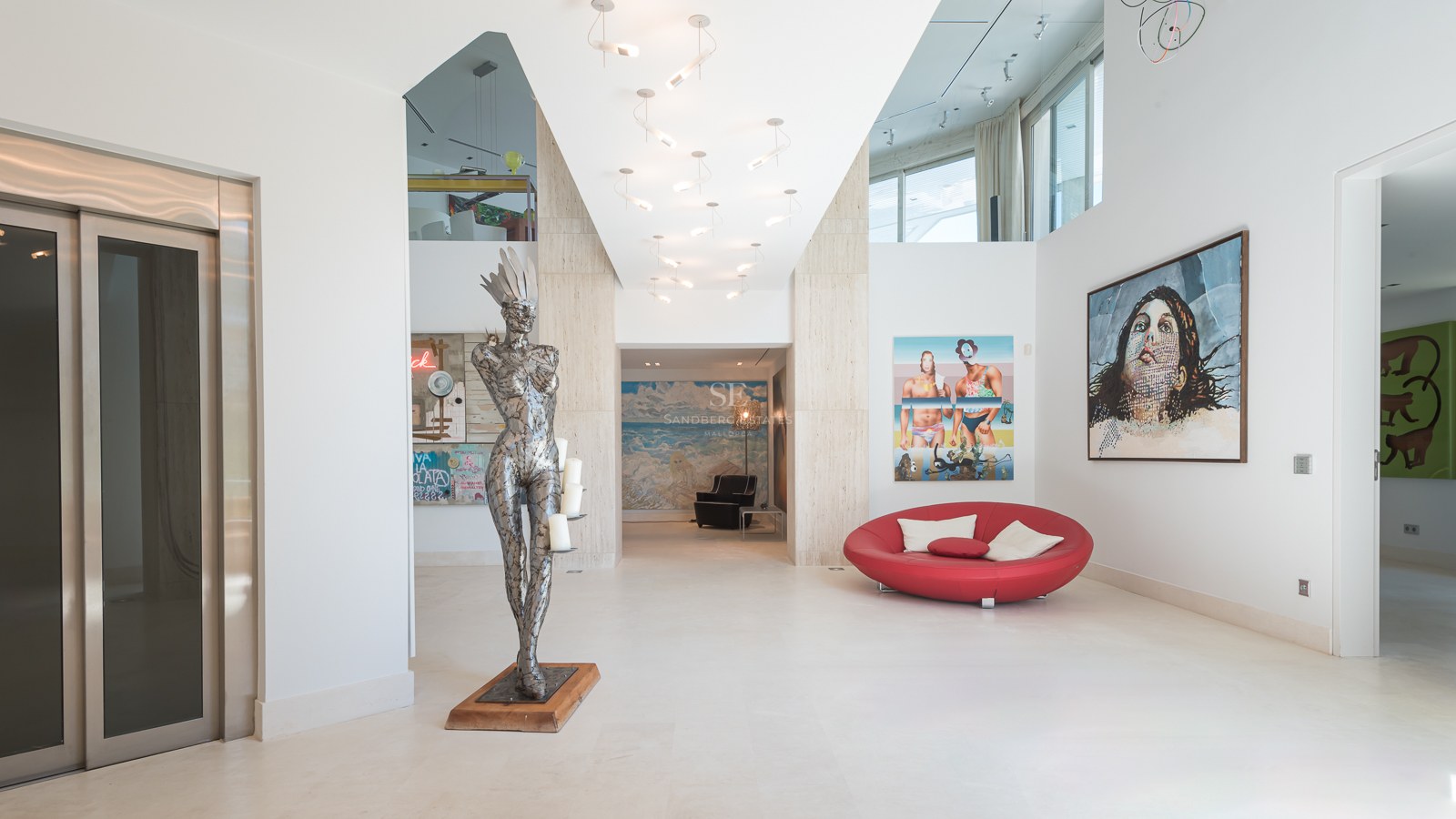 Modern white entrance hall with a private elevator, metal sculpture, red sofa, and large contemporary art pieces.