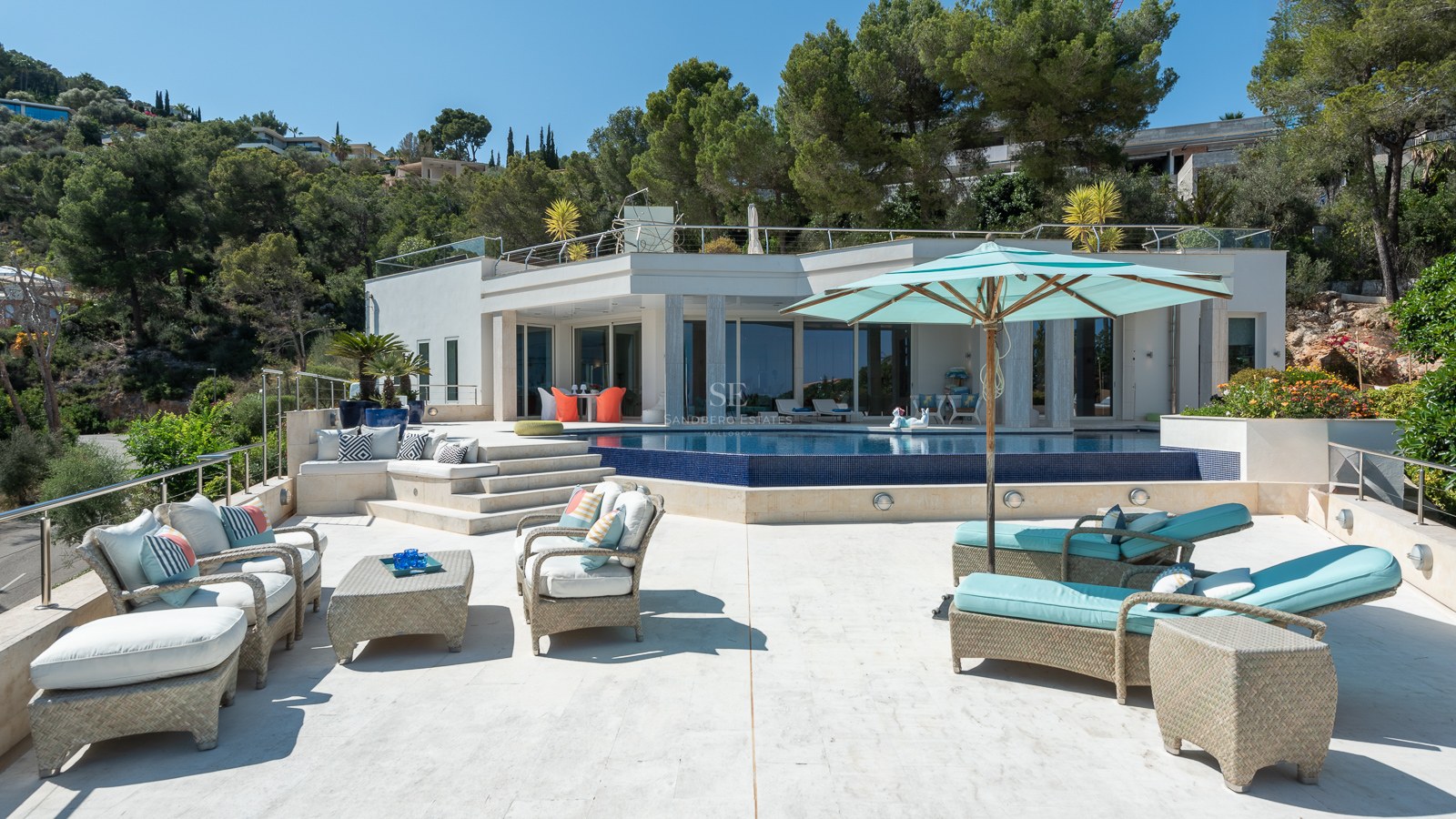 Outdoor terrace with turquoise sunbeds, wicker furniture, and a dark blue mosaic pool overlooking green hills.