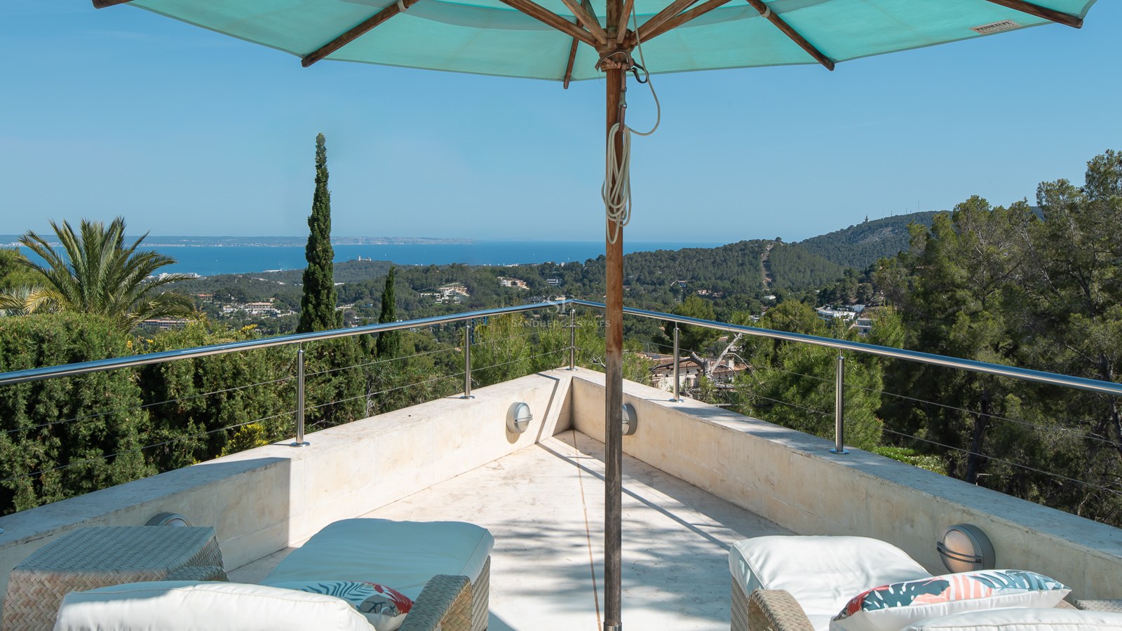 Elevated natural stone terrace with wicker loungers and turquoise parasol overlooking lush pine trees and the sea.