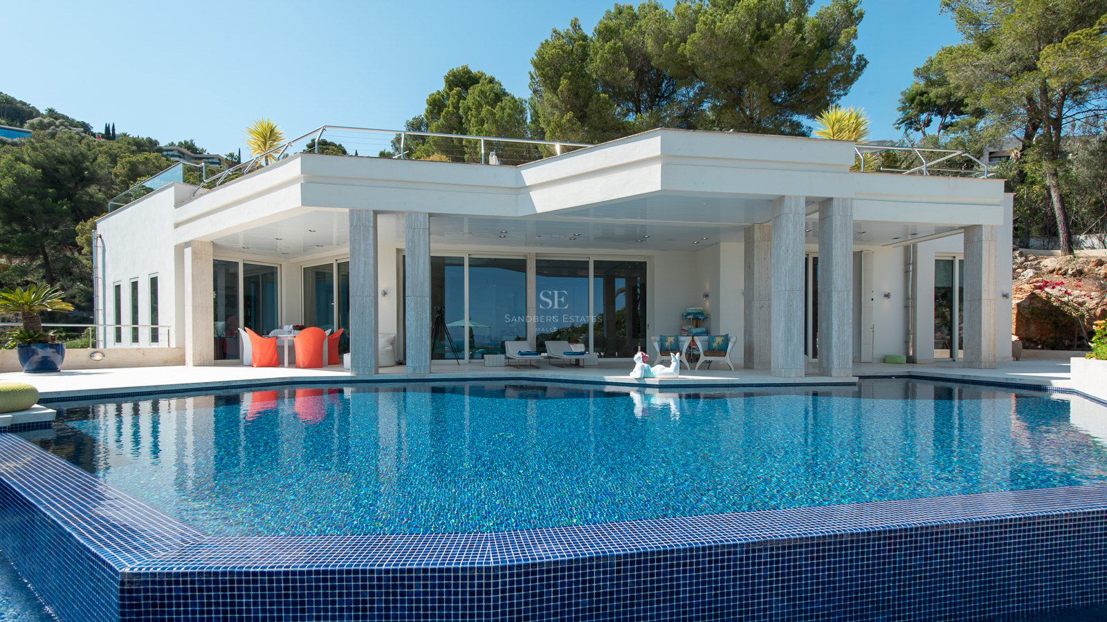 Modern white villa with a blue tiled swimming pool and a covered terrace with marble pillars surrounded by pine trees.