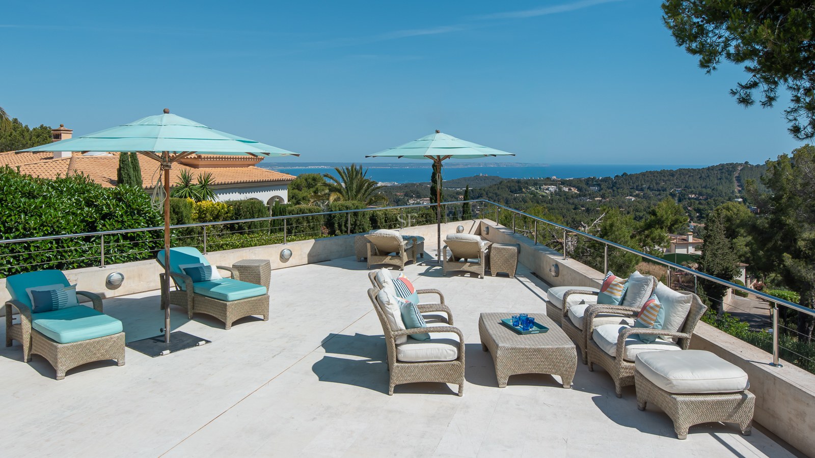 Elevated stone terrace with wicker lounge furniture and teal umbrellas overlooking a pine forest and the sea.