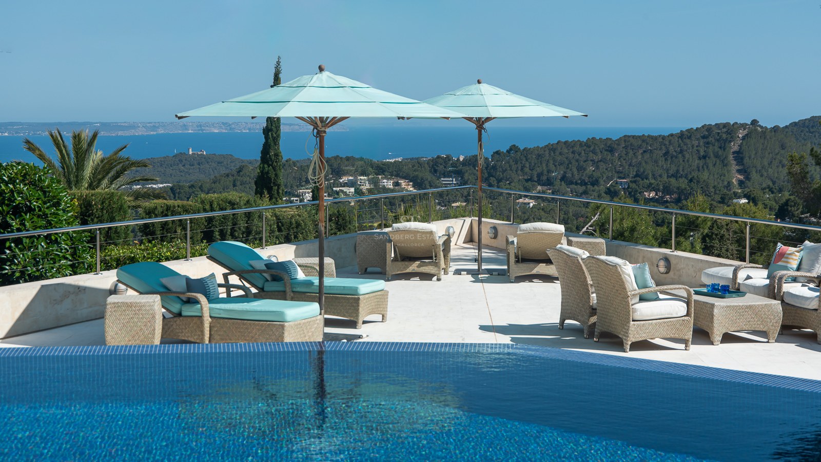 Infinity pool edge overlooking a large stone terrace with wicker furniture and light blue parasols facing the sea.