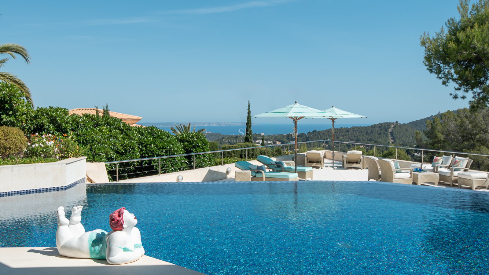 An infinity pool with blue mosaic tiles overlooking the Mediterranean coastline, featuring lounge chairs and a sculpture.