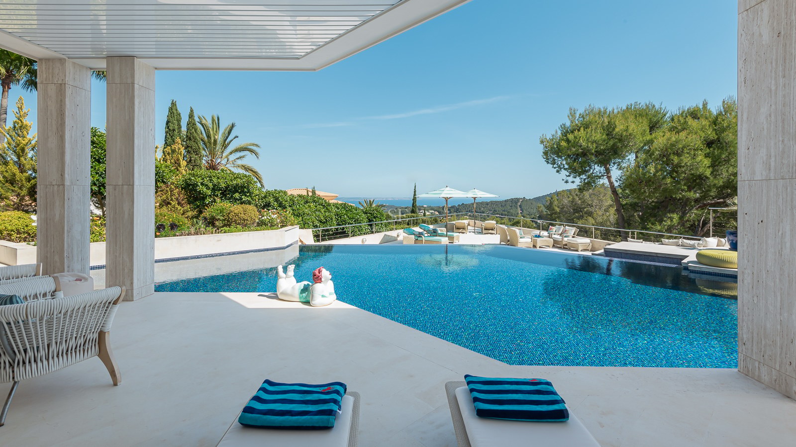 Luxury stone terrace featuring an azure infinity pool, lounge chairs, and panoramic sea views under a clear sky.