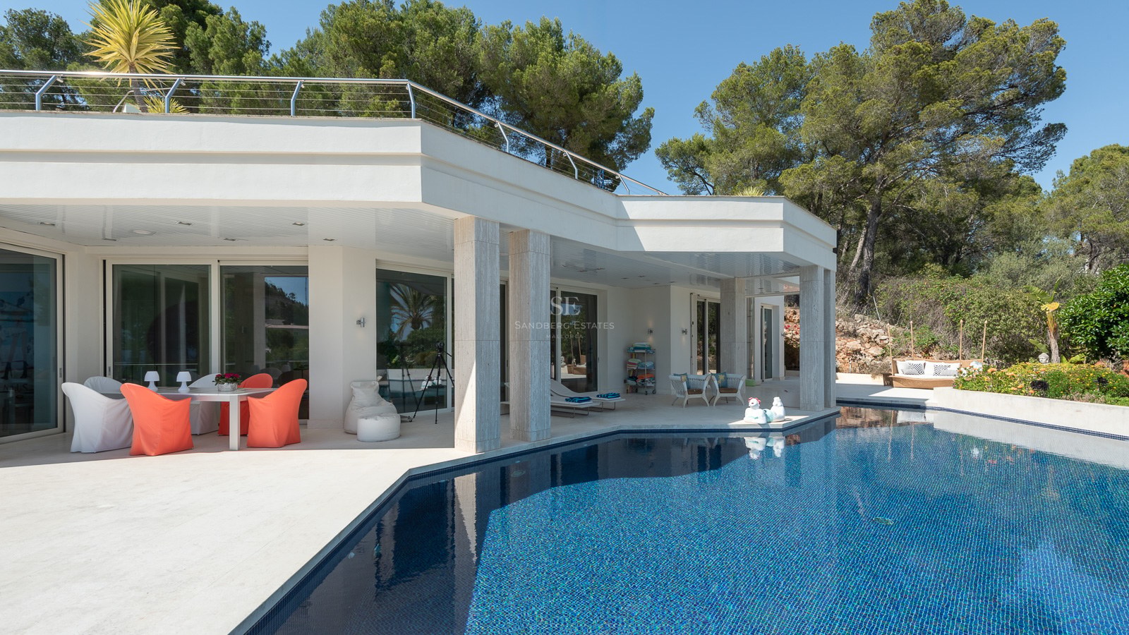 Exterior view of a white modern villa featuring a large blue swimming pool and shaded outdoor dining area.