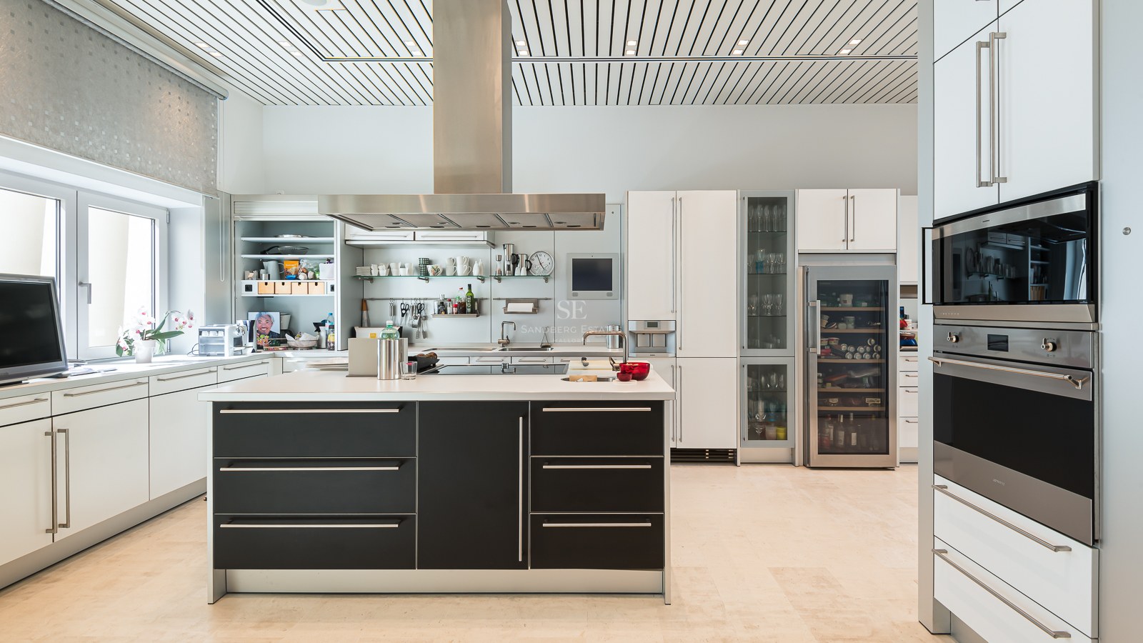 Modern white cabinetry with a black central island, stainless steel appliances, wine fridge, and light stone flooring.