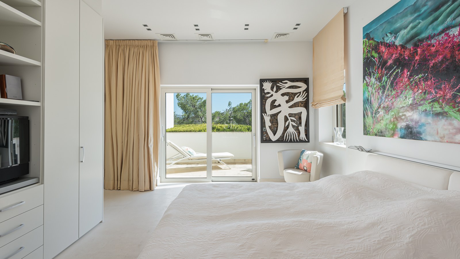 Bright modern bedroom featuring white walls, sliding glass doors to a terrace with sunbed, and colorful contemporary art.