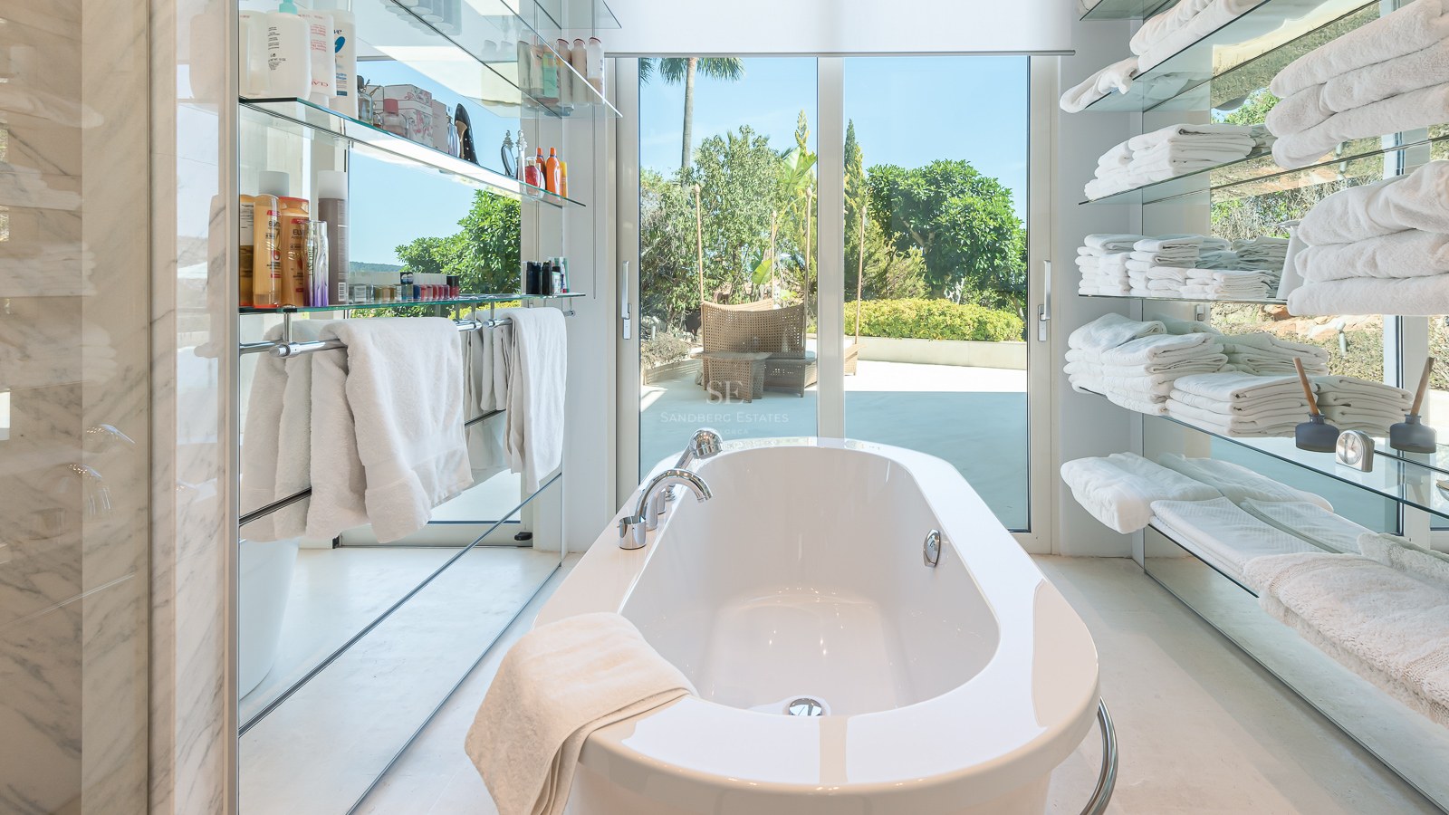 White freestanding bathtub centered between glass shelves, looking out through glass doors to a sunny garden.