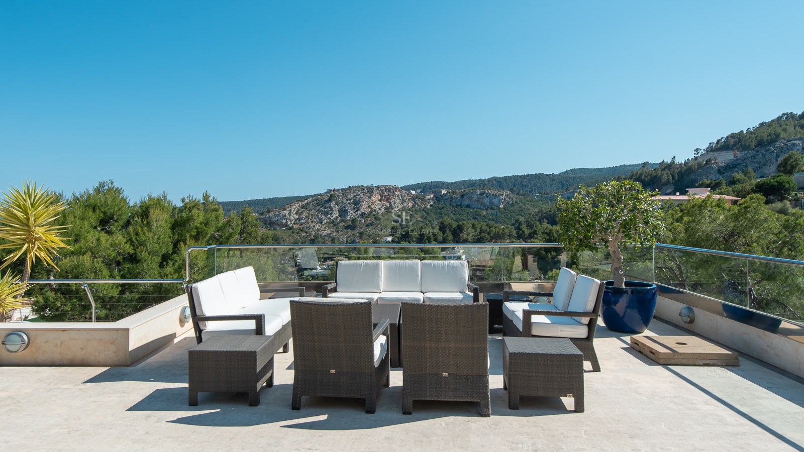 Spacious stone terrace with wicker seating area overlooking a lush mountain landscape under a clear blue sky.