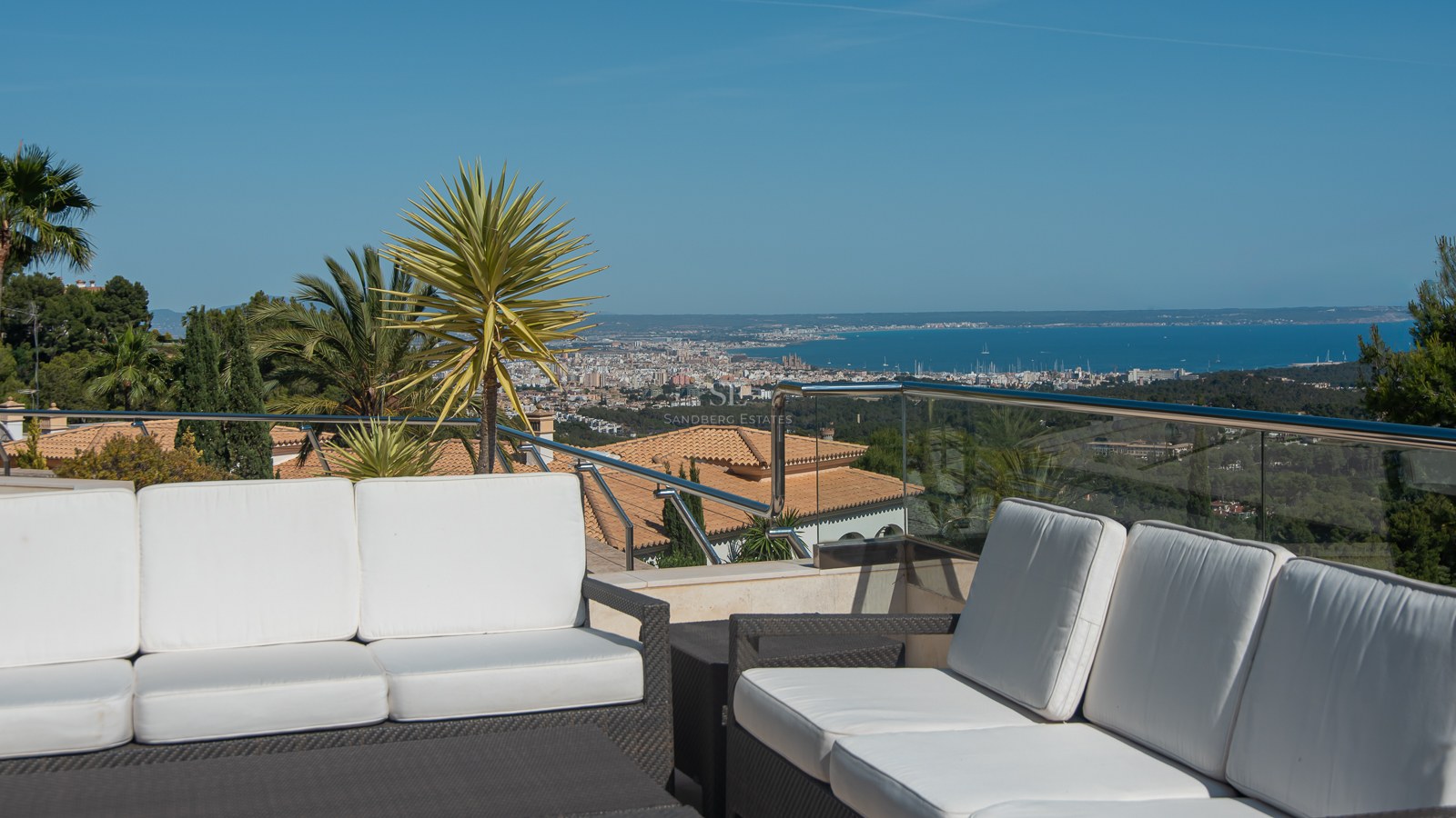 Luxury terrace with white sofas and glass railing overlooking a coastal city and the sea under a clear blue sky.