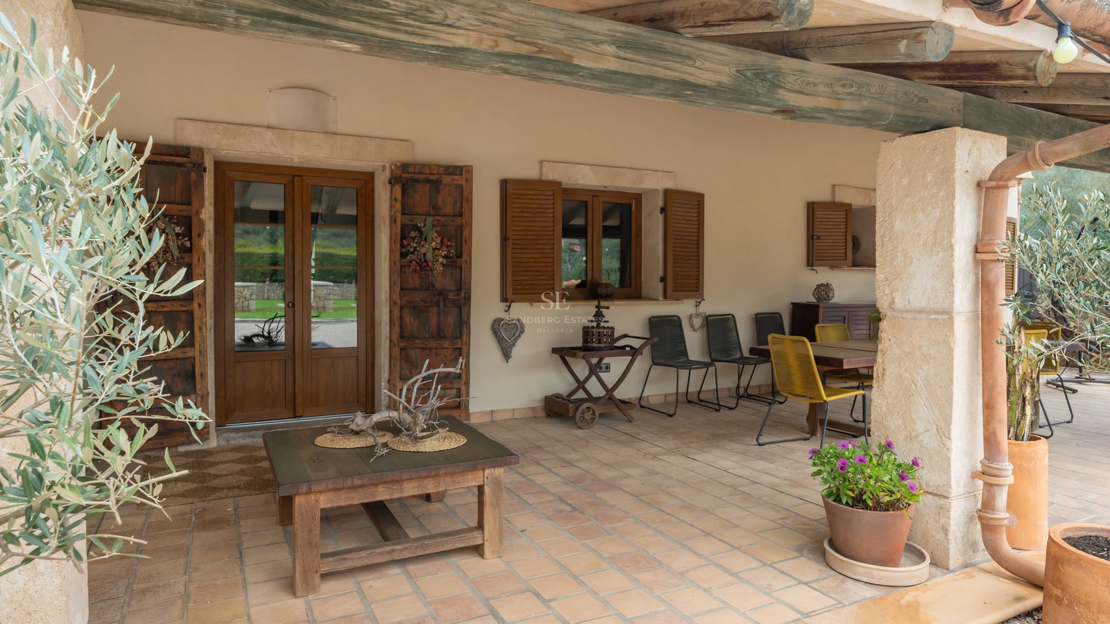 Porche couvert avec carrelage en terre cuite, poutres en bois rustiques, coin repas et murs en pierre claire.
