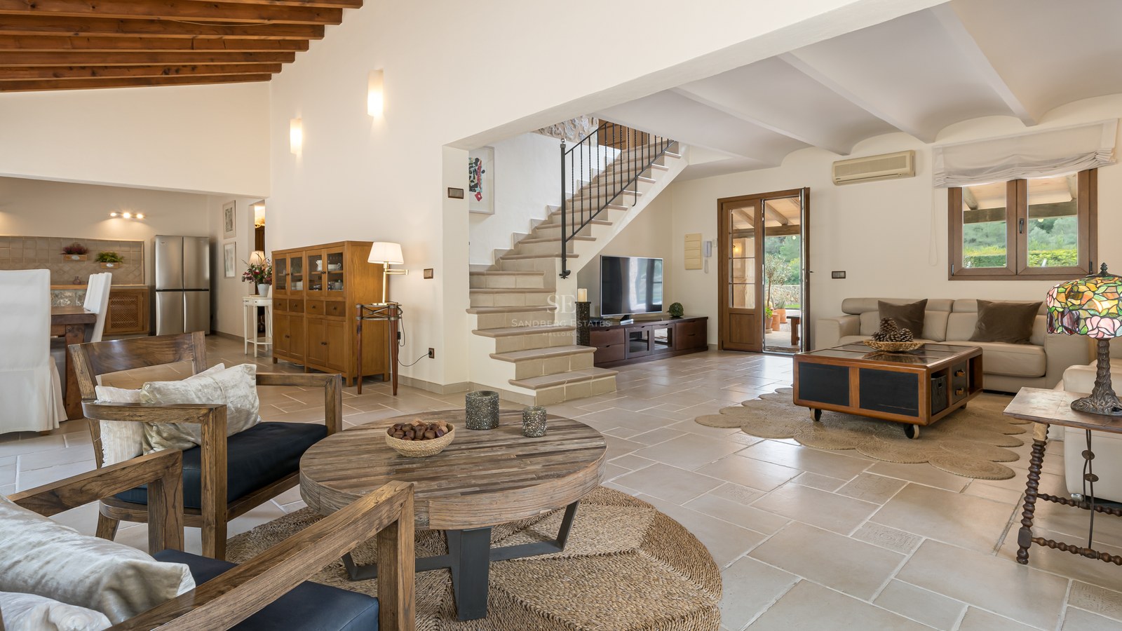 Grand salon avec sol en pierre naturelle, poutres en bois au plafond, escalier en pierre et mobilier classique.