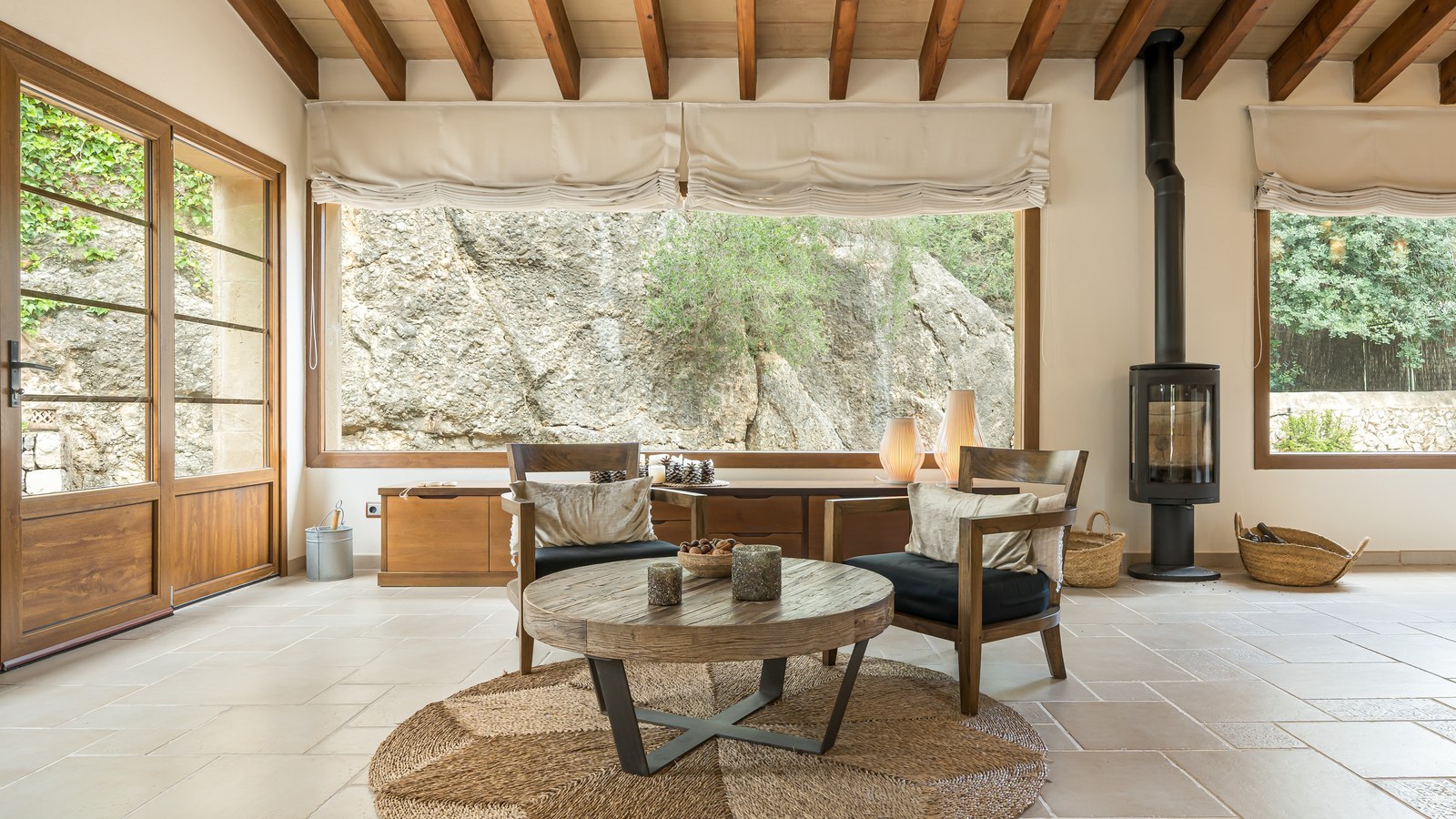 Salon lumineux avec poutres en bois, poêle à bois moderne et vue sur une paroi rocheuse naturelle.