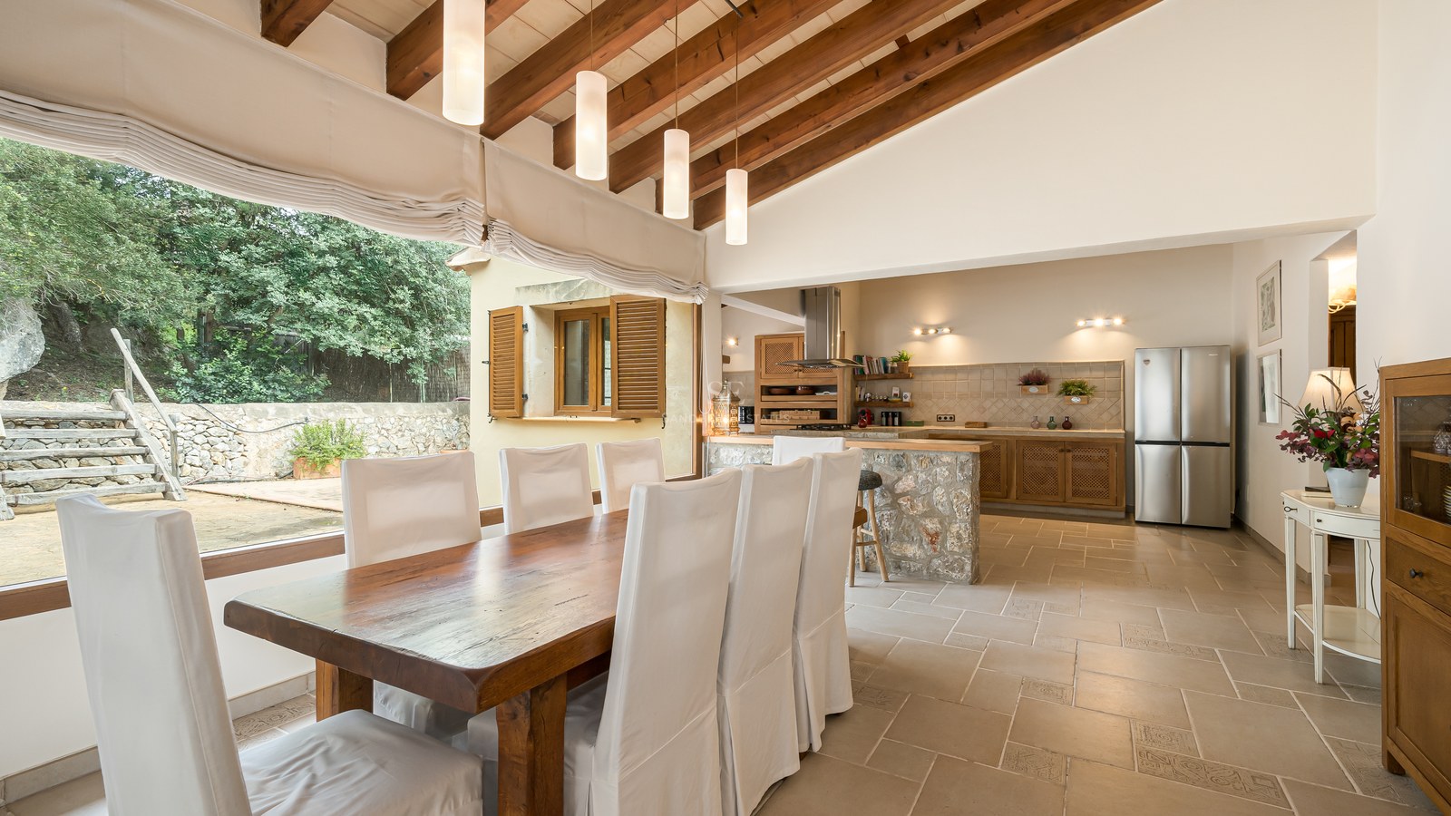 Salle à manger spacieuse avec poutres apparentes, sol en pierre et vue sur le jardin à travers de grandes fenêtres.