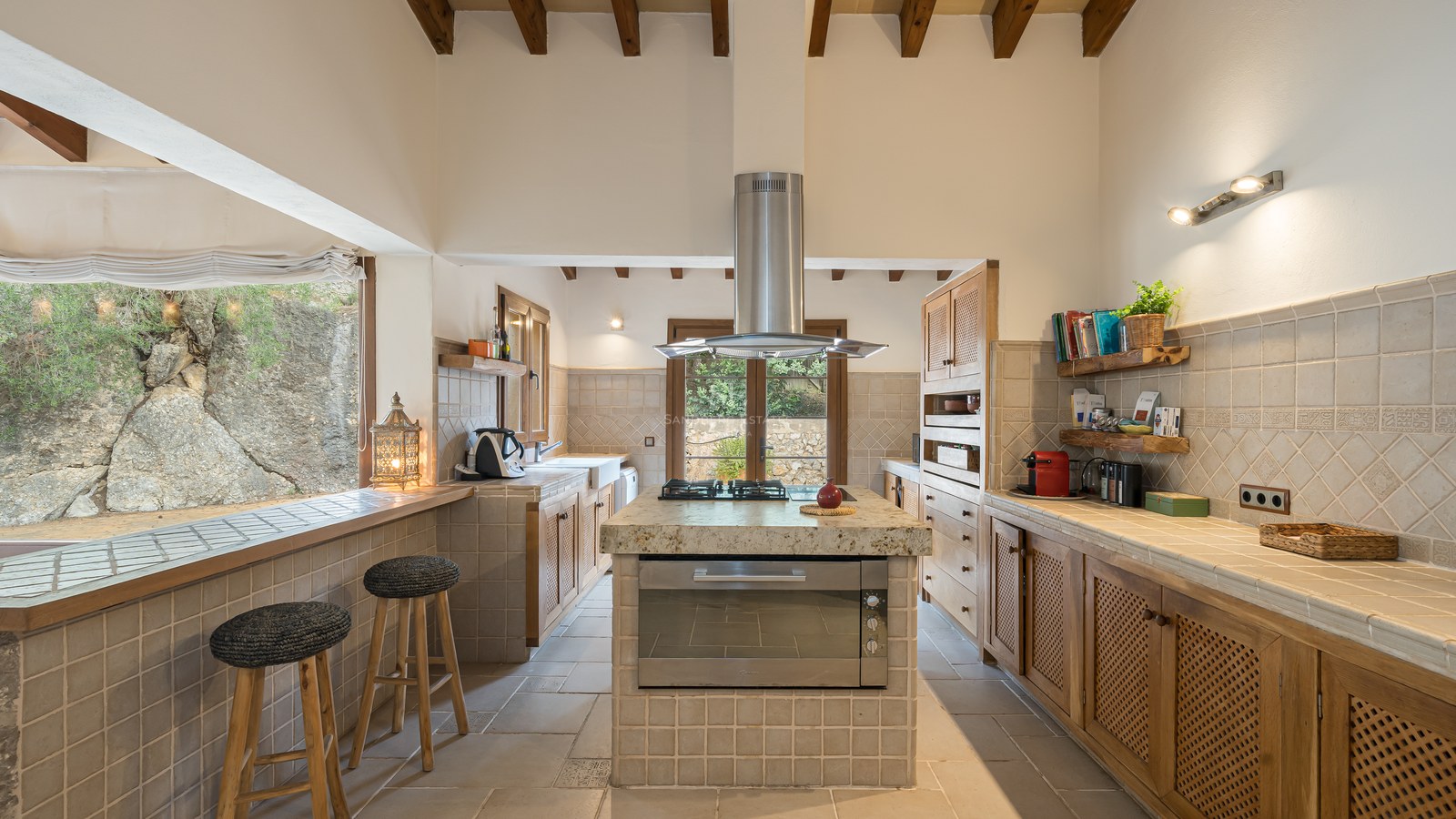 Cuisine rustique avec poutres apparentes, îlot central, plans de travail carrelés et taburets près d'une fenêtre.