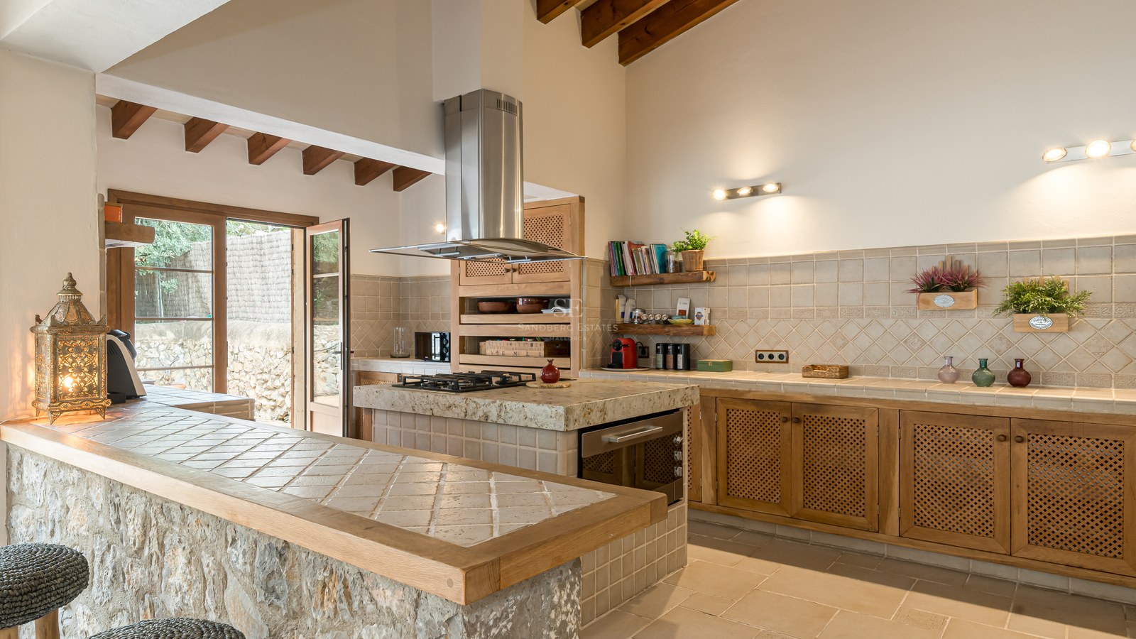 Grande cuisine avec poutres en bois, comptoir en pierre naturelle et îlot carrelé avec plaques de cuisson.