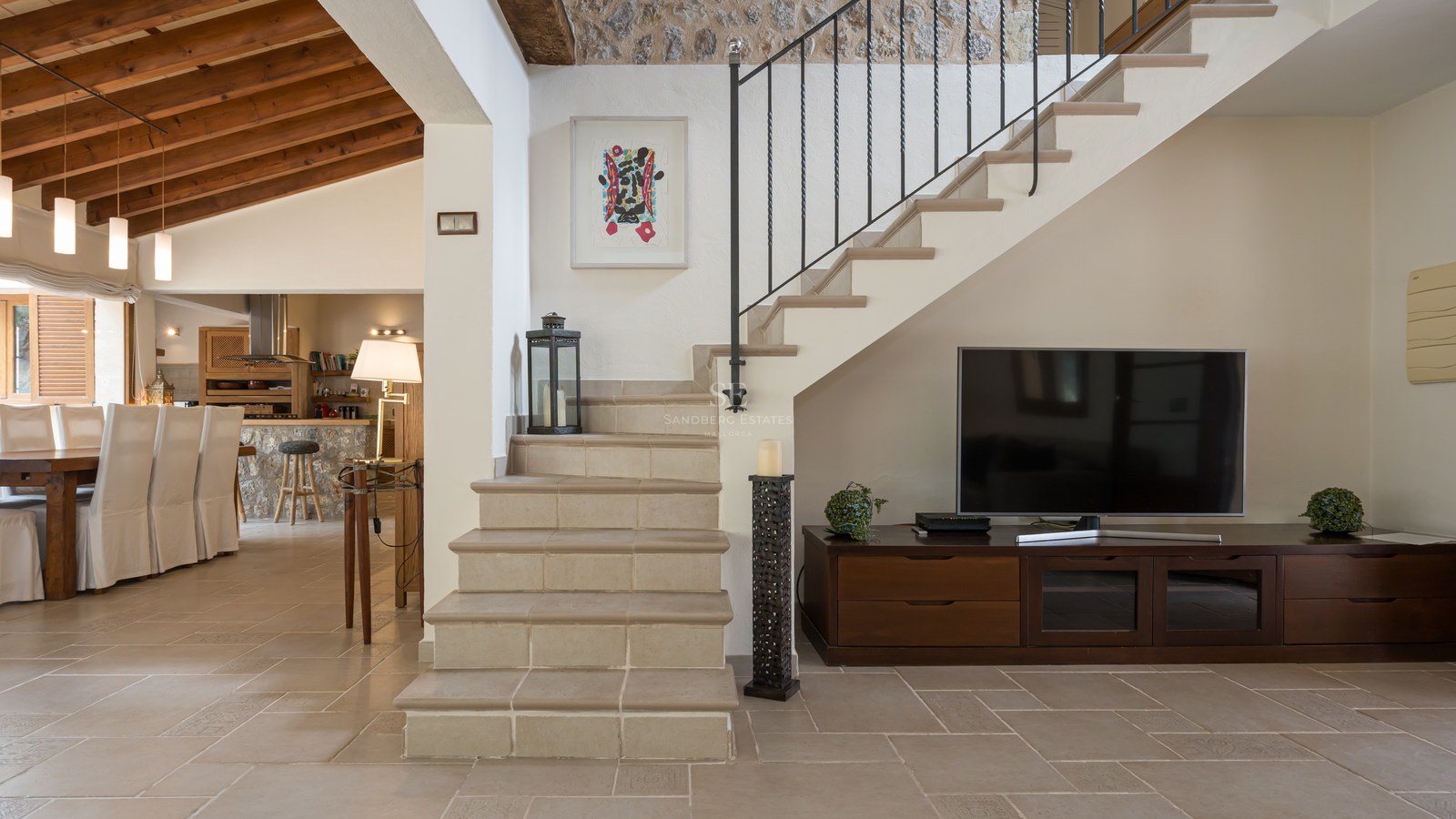 Vue intérieure montrant un escalier en pierre, un meuble TV en bois et un coin repas avec des poutres apparentes.