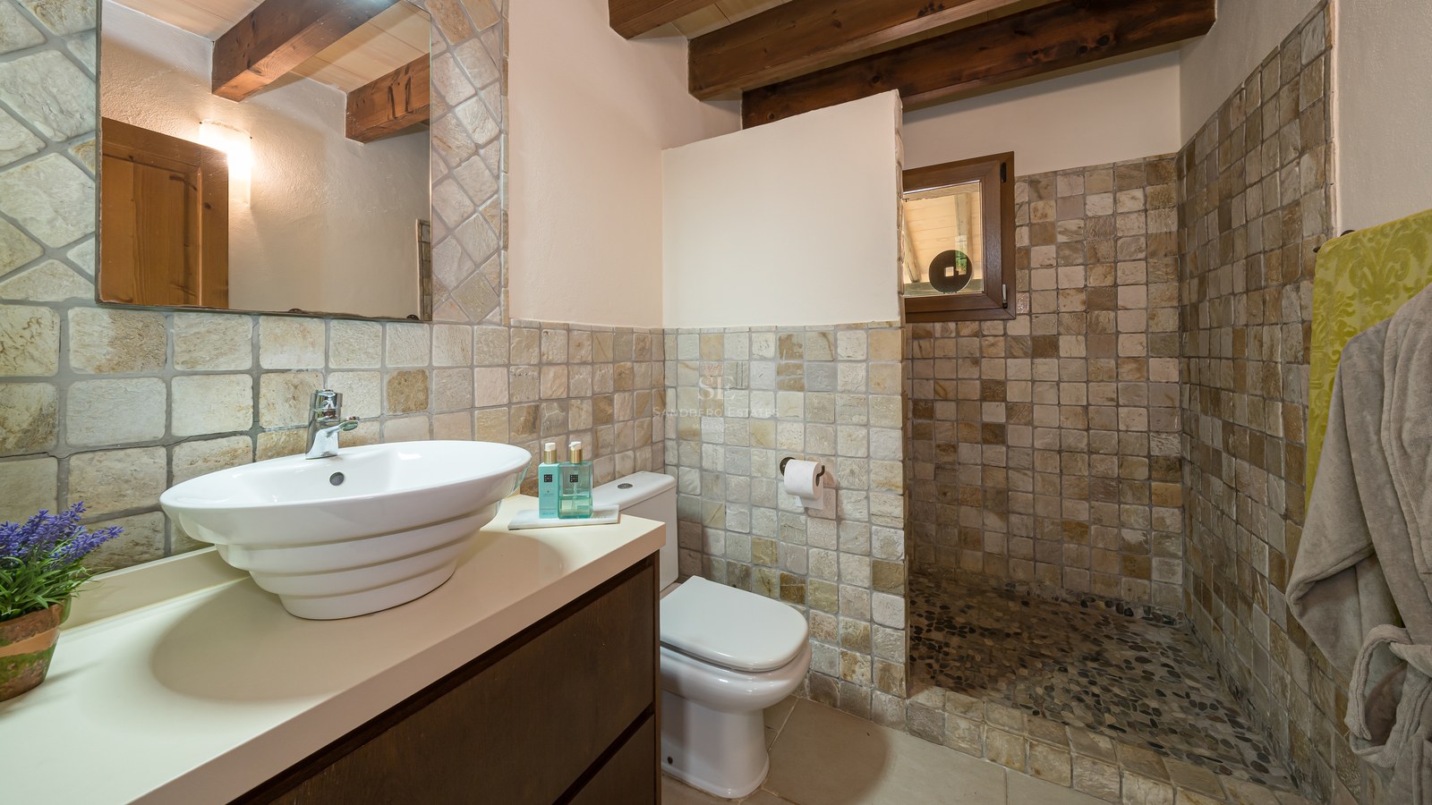 Salle de bain rustique avec murs en pierre, poutres apparentes, vasque blanche et douche au sol en galets.