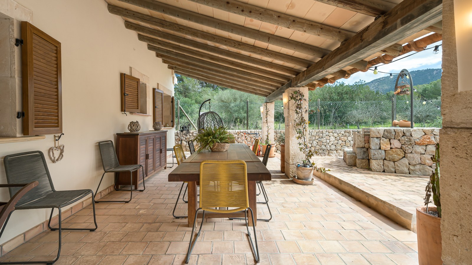 Terrasse couverte avec table en bois, chaises, murs en pierre et plafond à poutres avec vue sur la campagne.