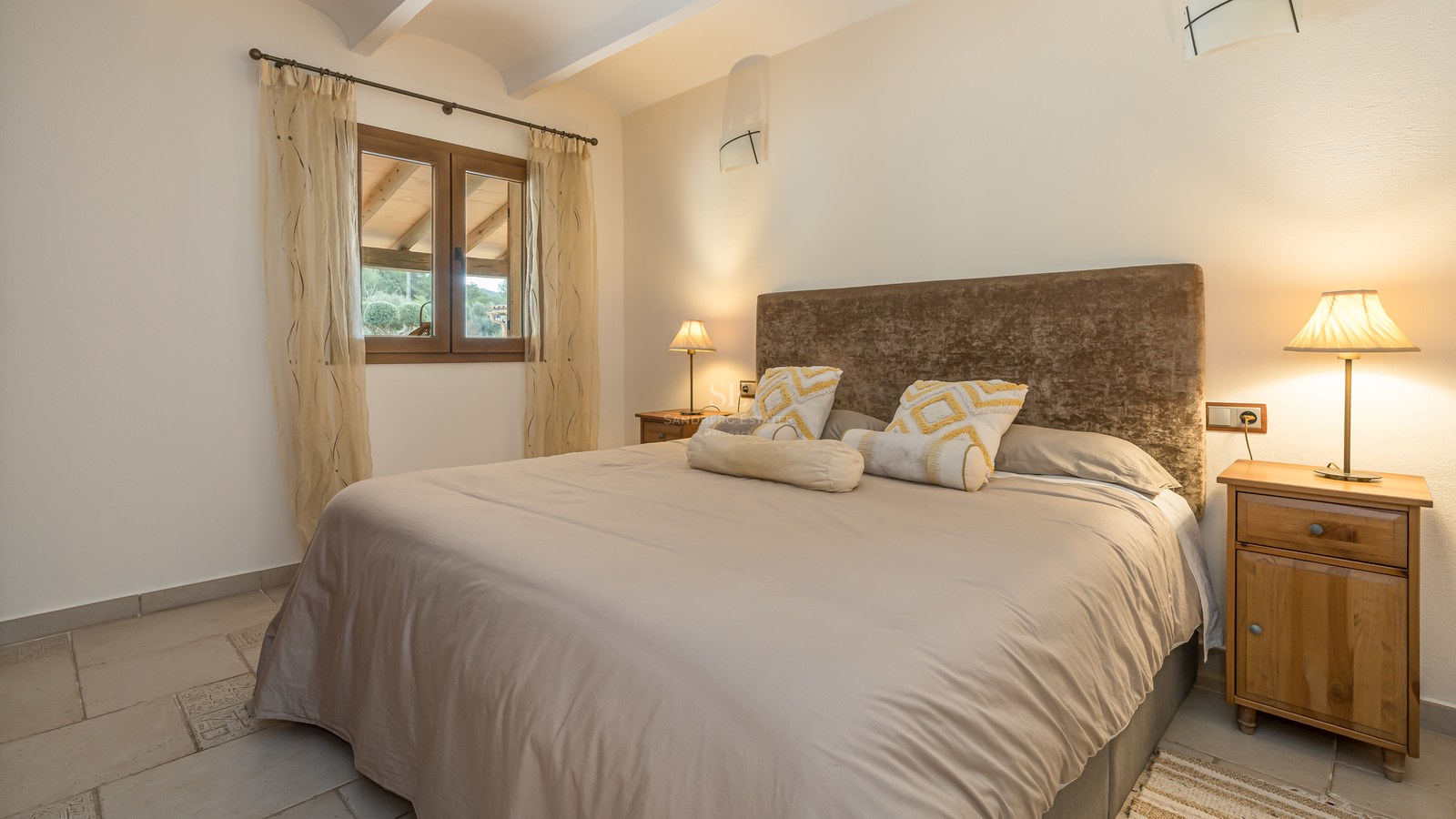 Chambre chaleureuse avec grand lit, tête de lit en velours marron, tables de chevet en bois et éclairage tamisé.
