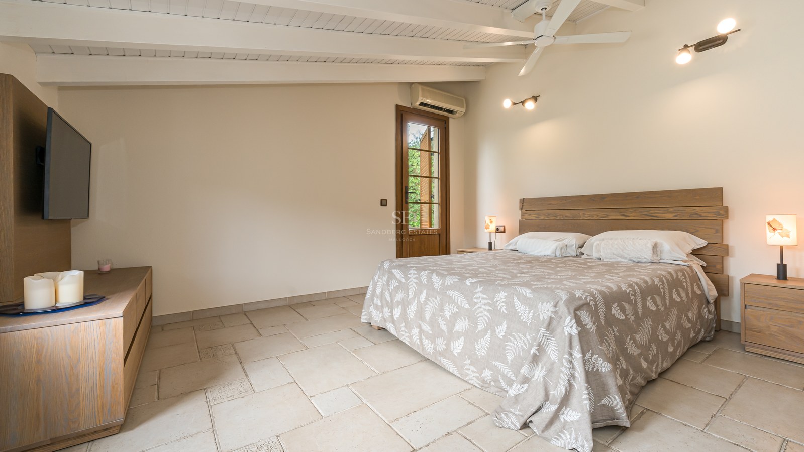 Chambre avec lit en bois, plafond à poutres blanches, sol en pierre et grandes fenêtres.