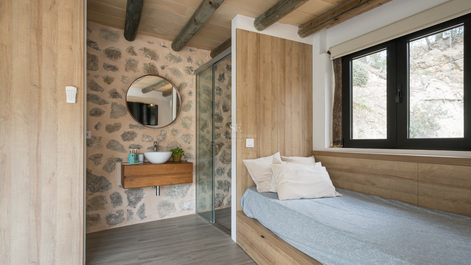 Chambre avec mur en pierre apparente, poutres en bois, lit intégré et coin vasque ouvert.