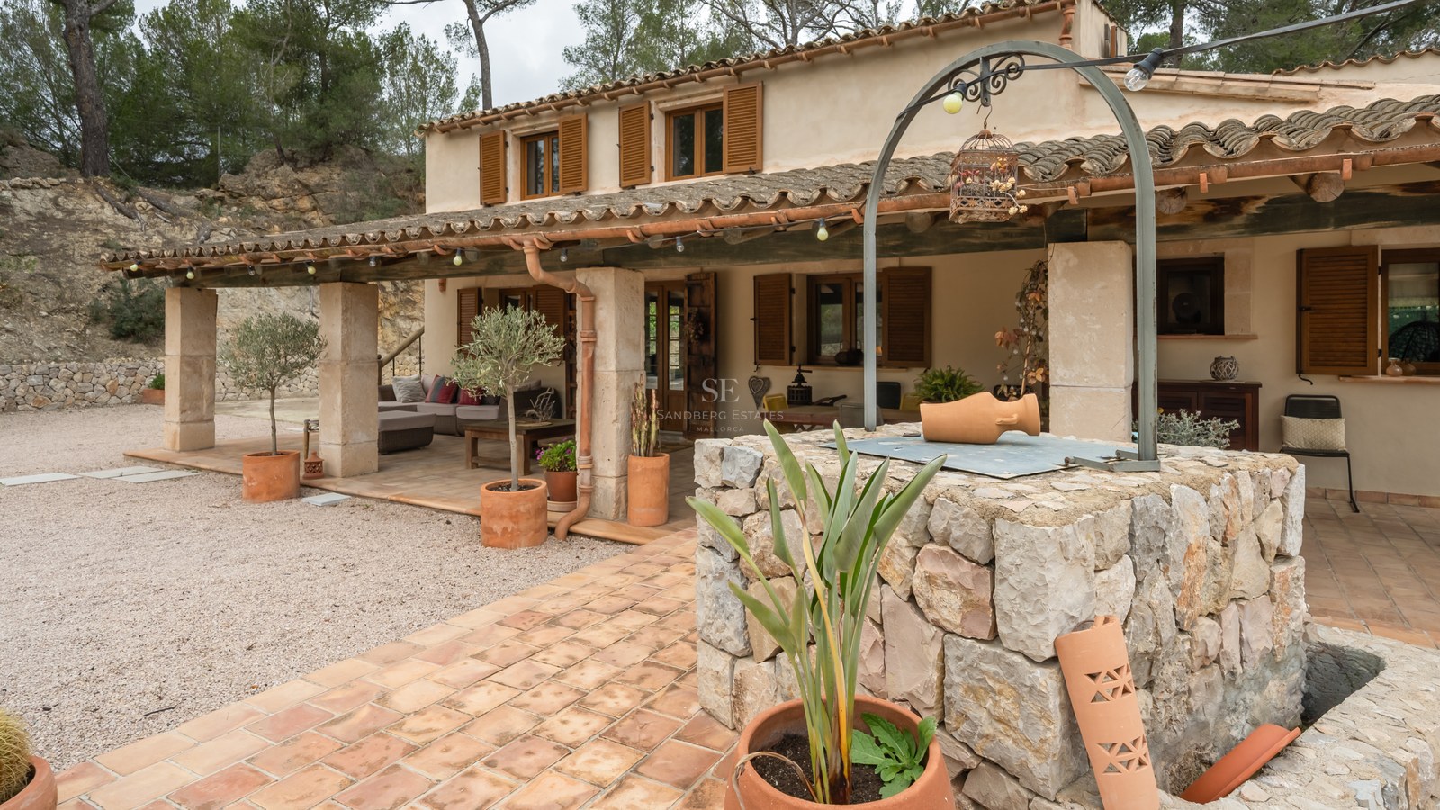 Façade d'une maison rustique avec porche couvert, volets en bois et un puits en pierre au premier plan.