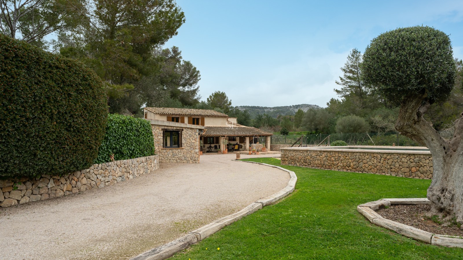 Maison en pierre rustique avec allée en gravier, pelouse verte et piscine en pierre surélevée sous un ciel clair.