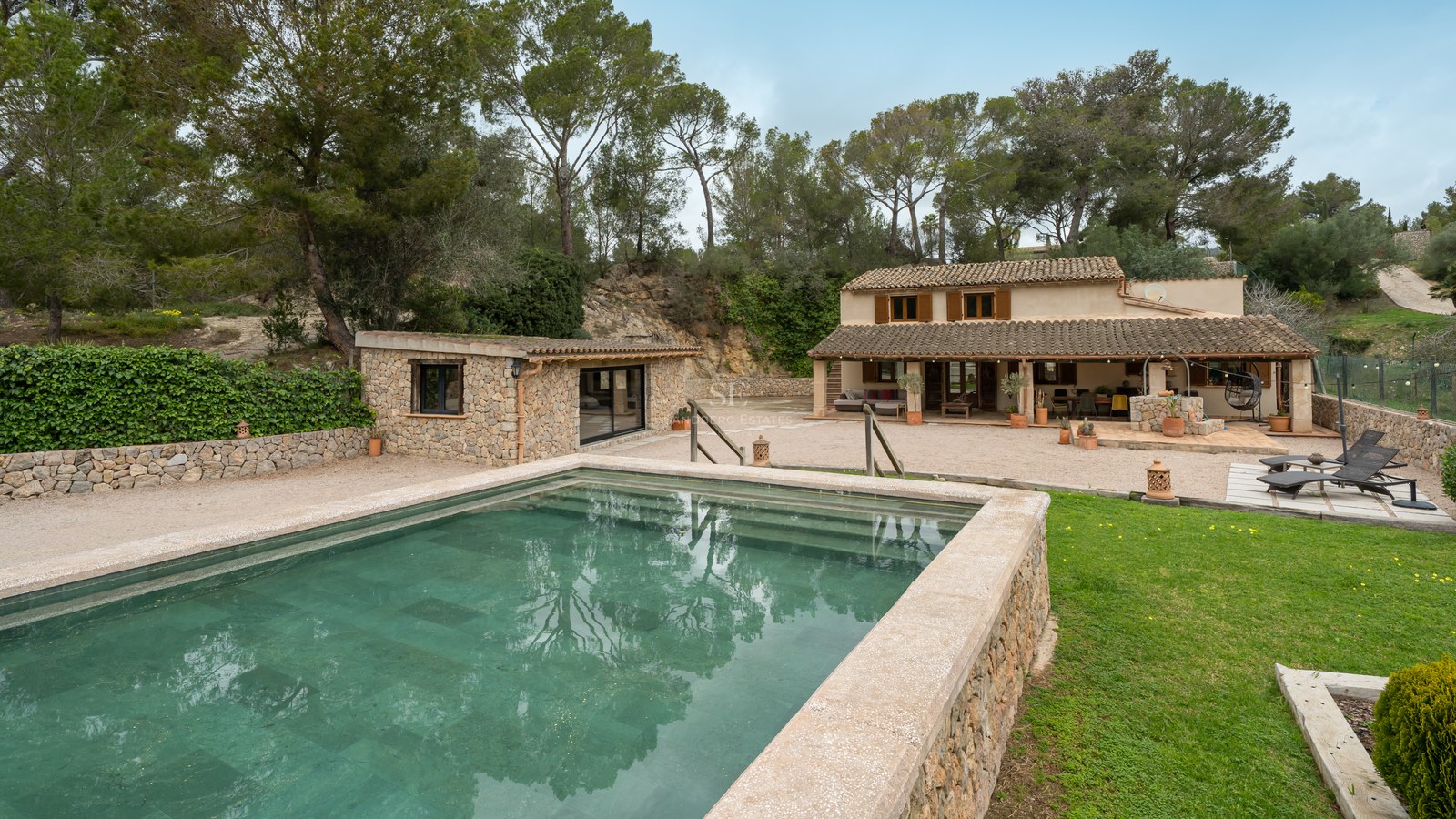 Piscine extérieure avec bordures en pierre devant une villa méditerranéenne rustique entourée de pins.