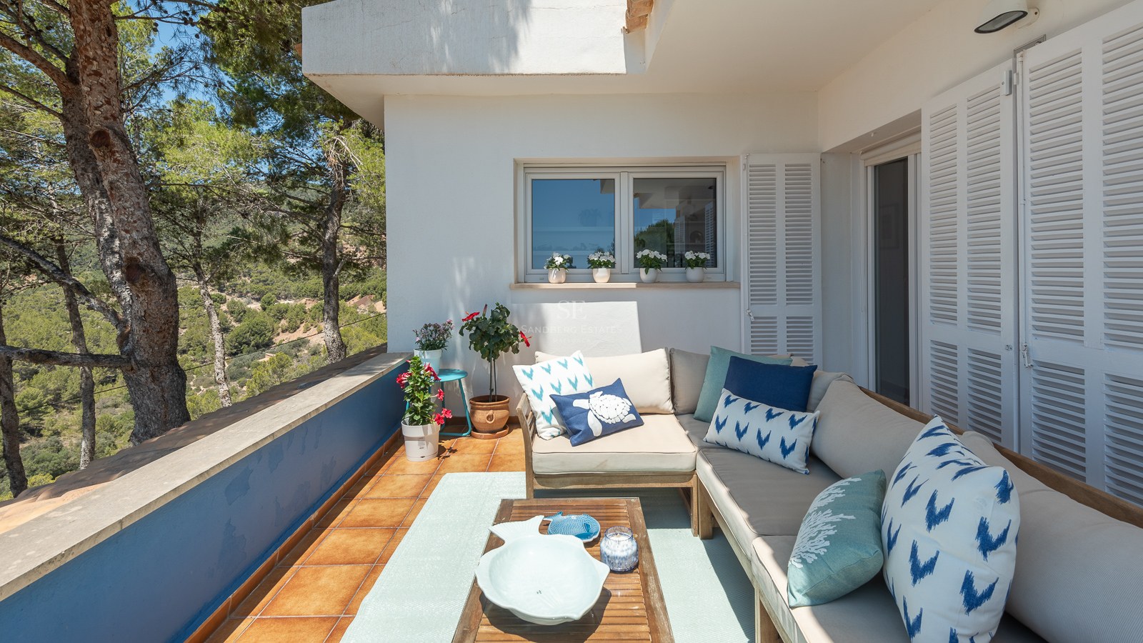 Terrasse extérieure avec un grand canapé en L, des coussins bleus et une table en bois surplombant une vallée de pins.