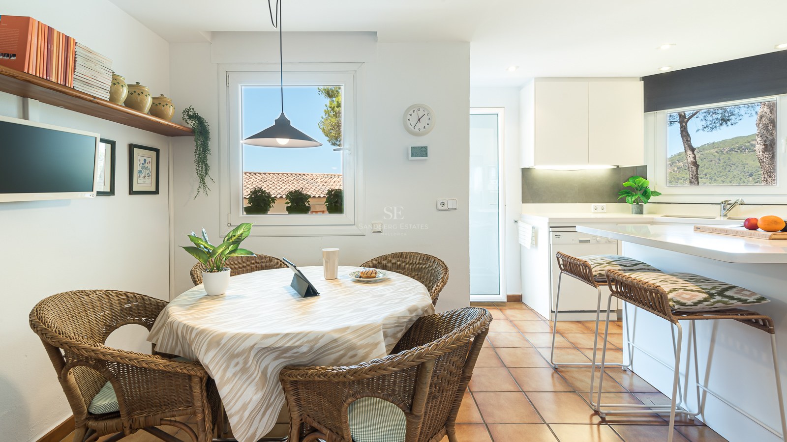 Table à manger ronde avec chaises en osier et sol en terre cuite à côté d'une cuisine moderne blanche.