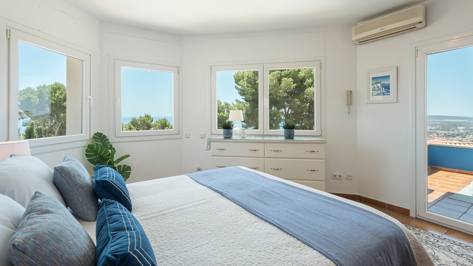 Chambre blanche lumineuse avec de grandes fenêtres vue mer, literie bleue et porte vers un balcon ensoleillé.