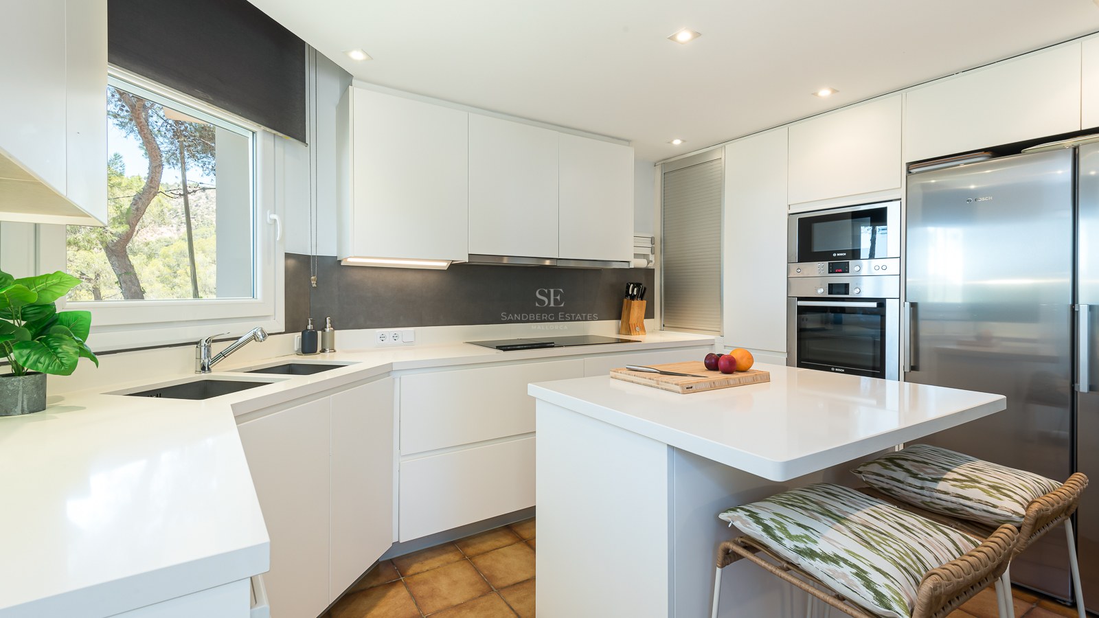 Cuisine moderne lumineuse avec placards blancs, appareils en inox, îlot central et vue sur la forêt par la fenêtre.