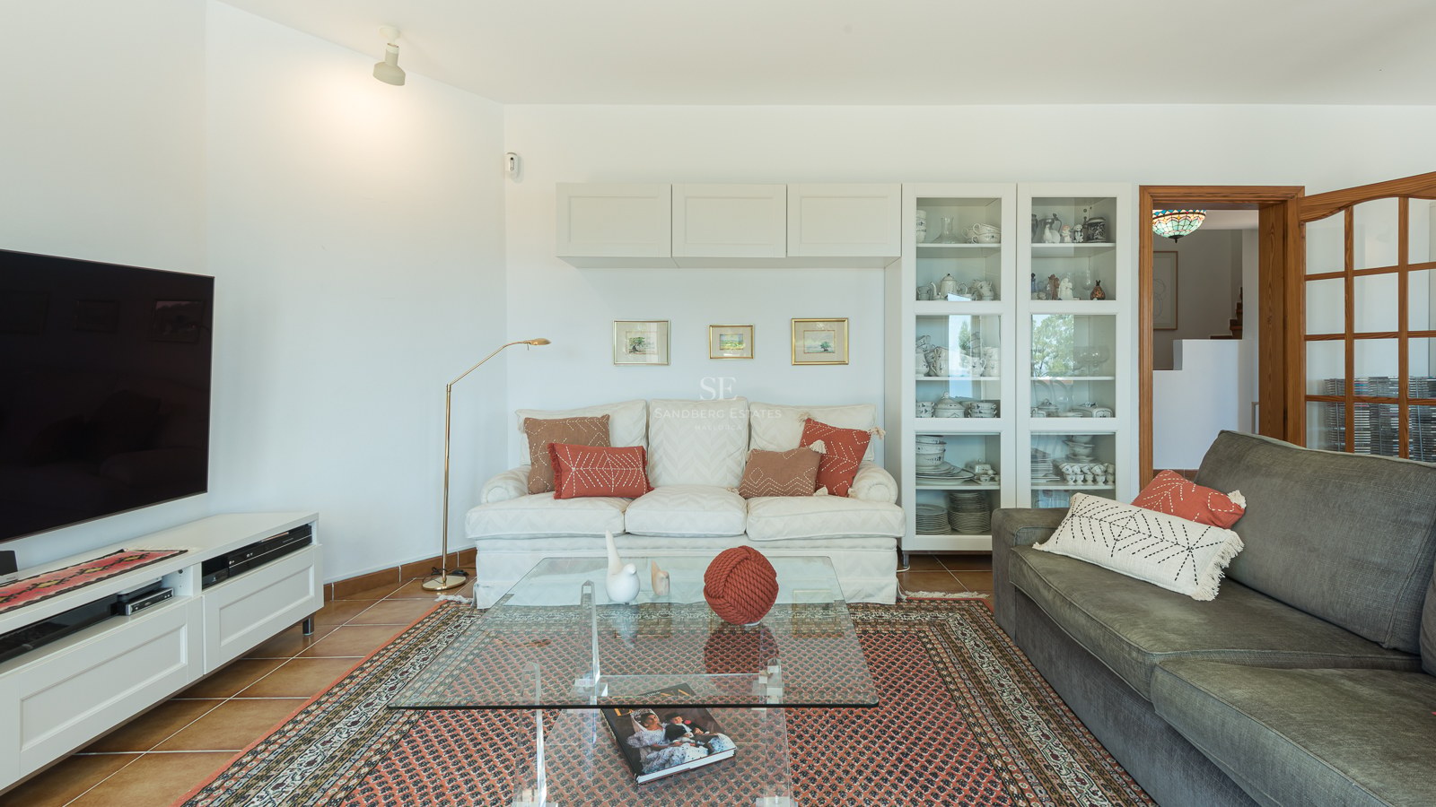 Salon lumineux avec canapés, table basse en verre sur tapis orné, grand téléviseur et sol en terre cuite.
