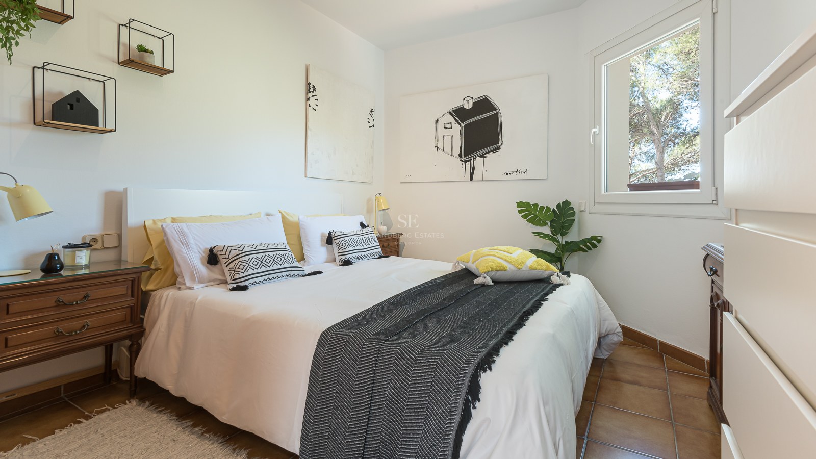 Chambre moderne avec lit blanc, coussins à motifs, tables de chevet en bois et sol en carrelage sous lumière naturelle.