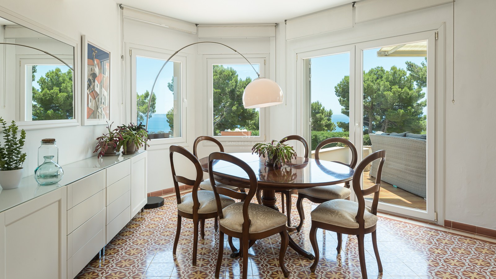 Salle à manger lumineuse avec table ronde en bois, sol en carreaux de ciment et grandes fenêtres donnant sur la mer.