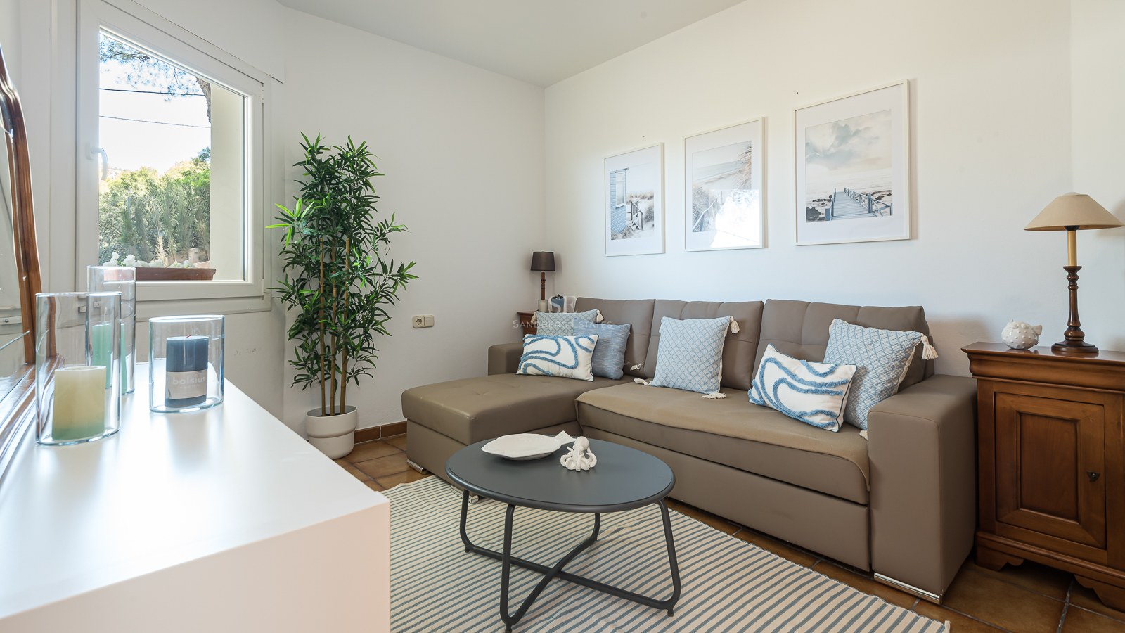 Un salon lumineux avec un canapé en L taupe, des coussins bleus, un tapis à rayures et des tableaux sur le thème de la mer.