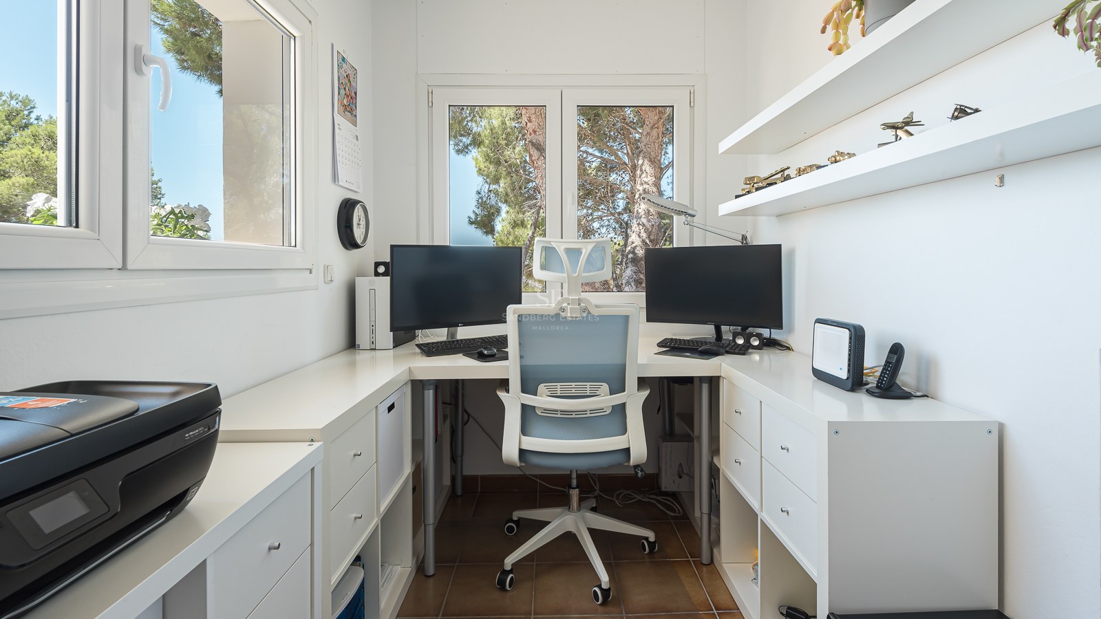 Bureau blanc moderne avec deux écrans, chaise ergonomique et fenêtres donnant sur des arbres verts.